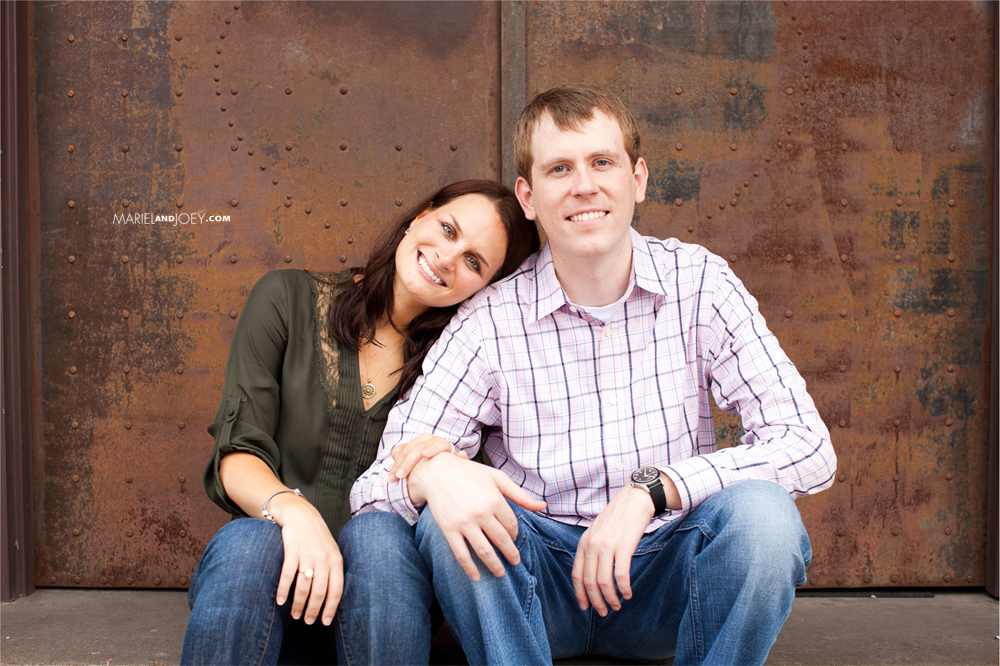 Houston couple sitting and smiling at camera in casual clothes in front of weathered door